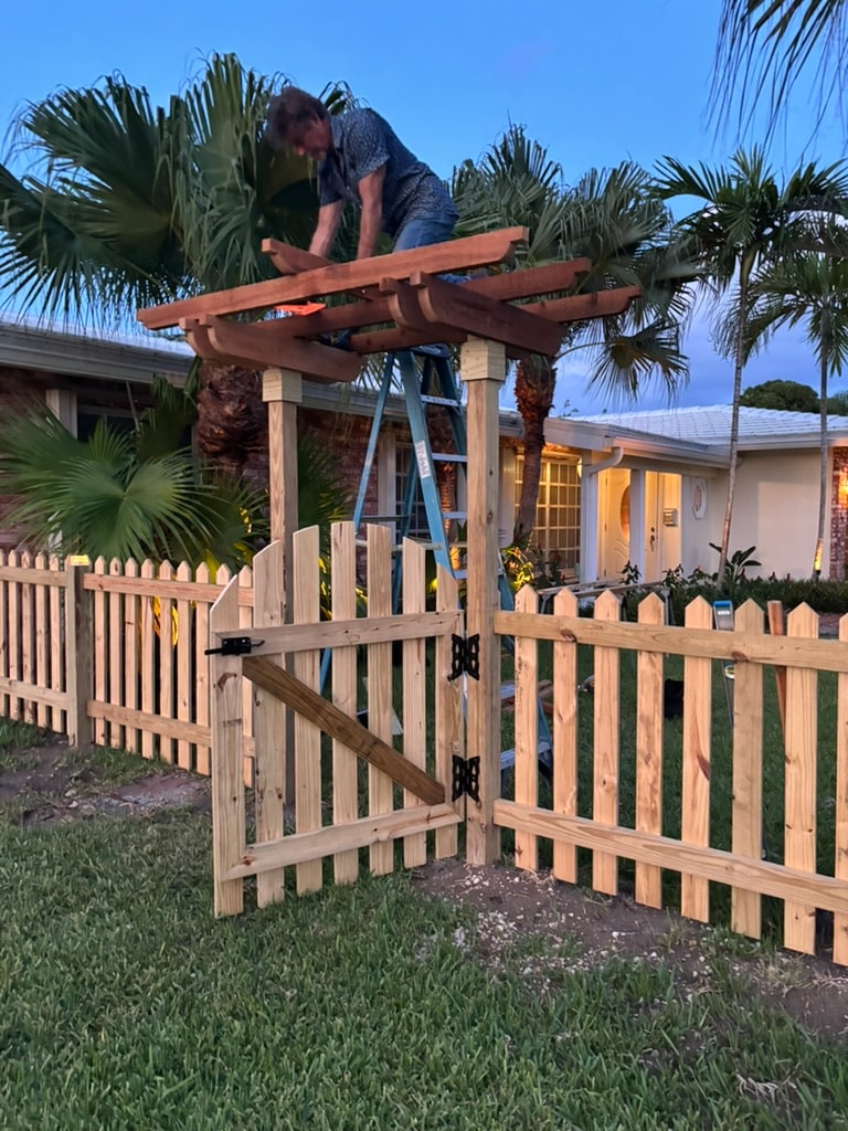 Worker finishing trellis installation over picket fence gate at dusk